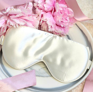 a gorgeous white organic mulberry silk sleep eye mask sits on a marble tray surrounded with luscious pink peonies.